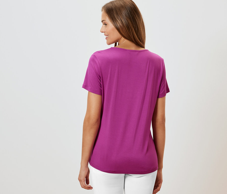 Hátulról látható nő bogyószínű, rakott részlettel ellátott pólóban és fehér nadrágban.