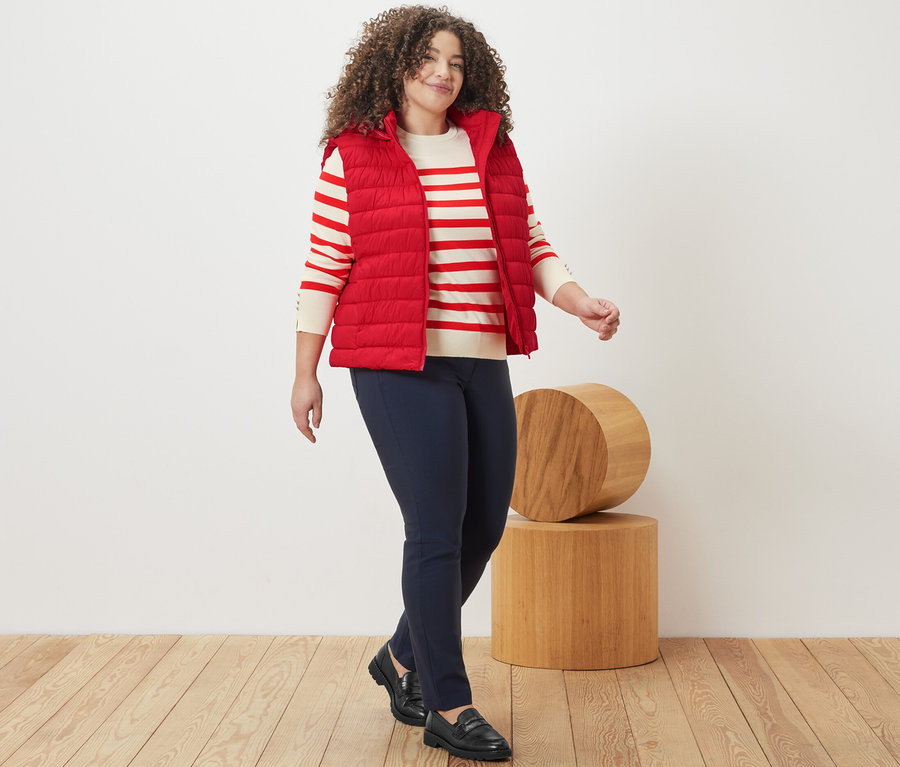 Nő sétál, piros csíkos finomkötésű pulóvert, kapucnis steppelt mellényt és sötétkék bengalin nadrágot visel.