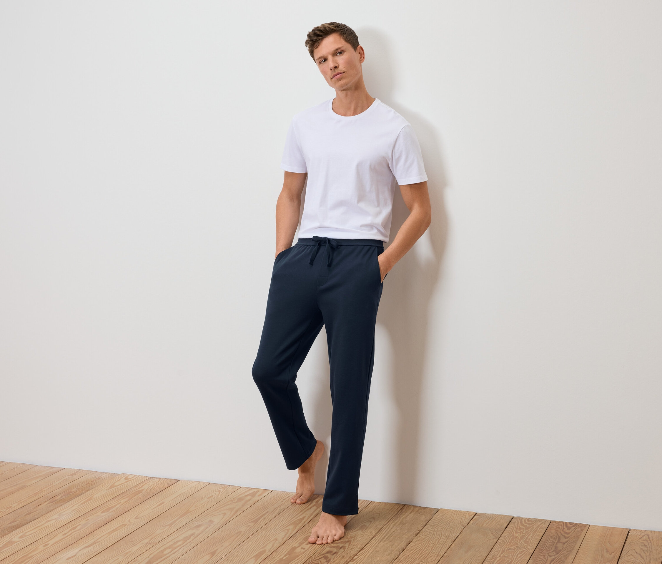 Férfi áll a falnak támaszkodva fehér pólóban és sötétkék melegítőnadrágban.