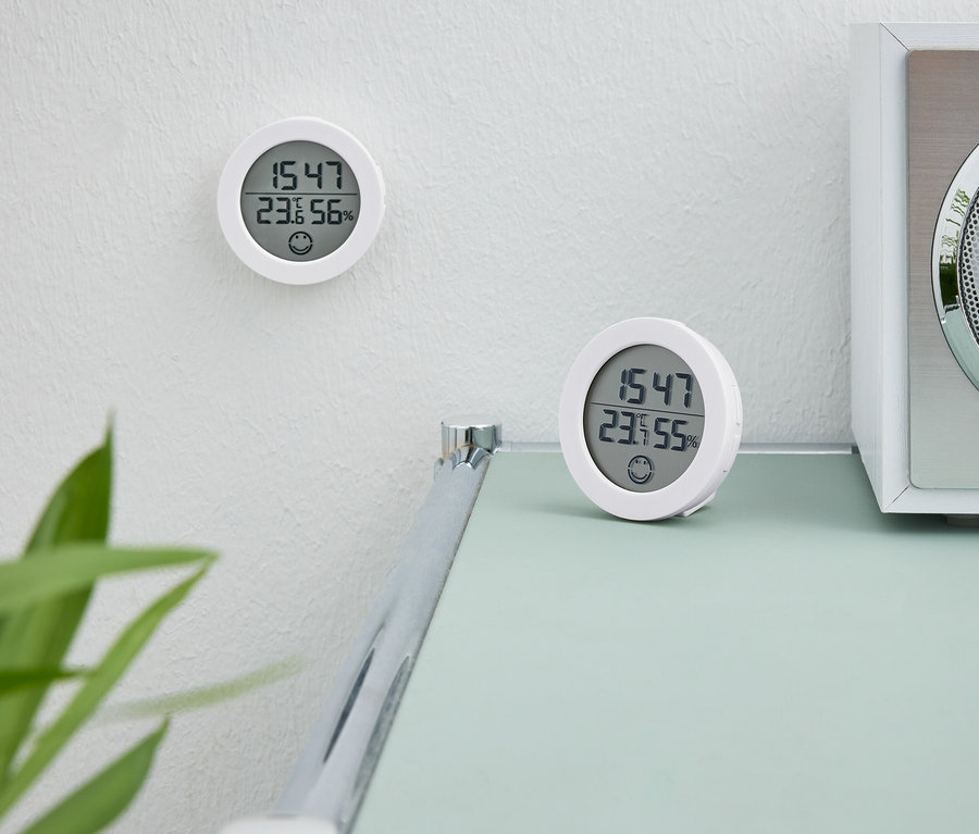 Két fehér digitális óra. Az egyik óra a falon van. A második óra egy asztalon áll egy fehér rádió mellett.