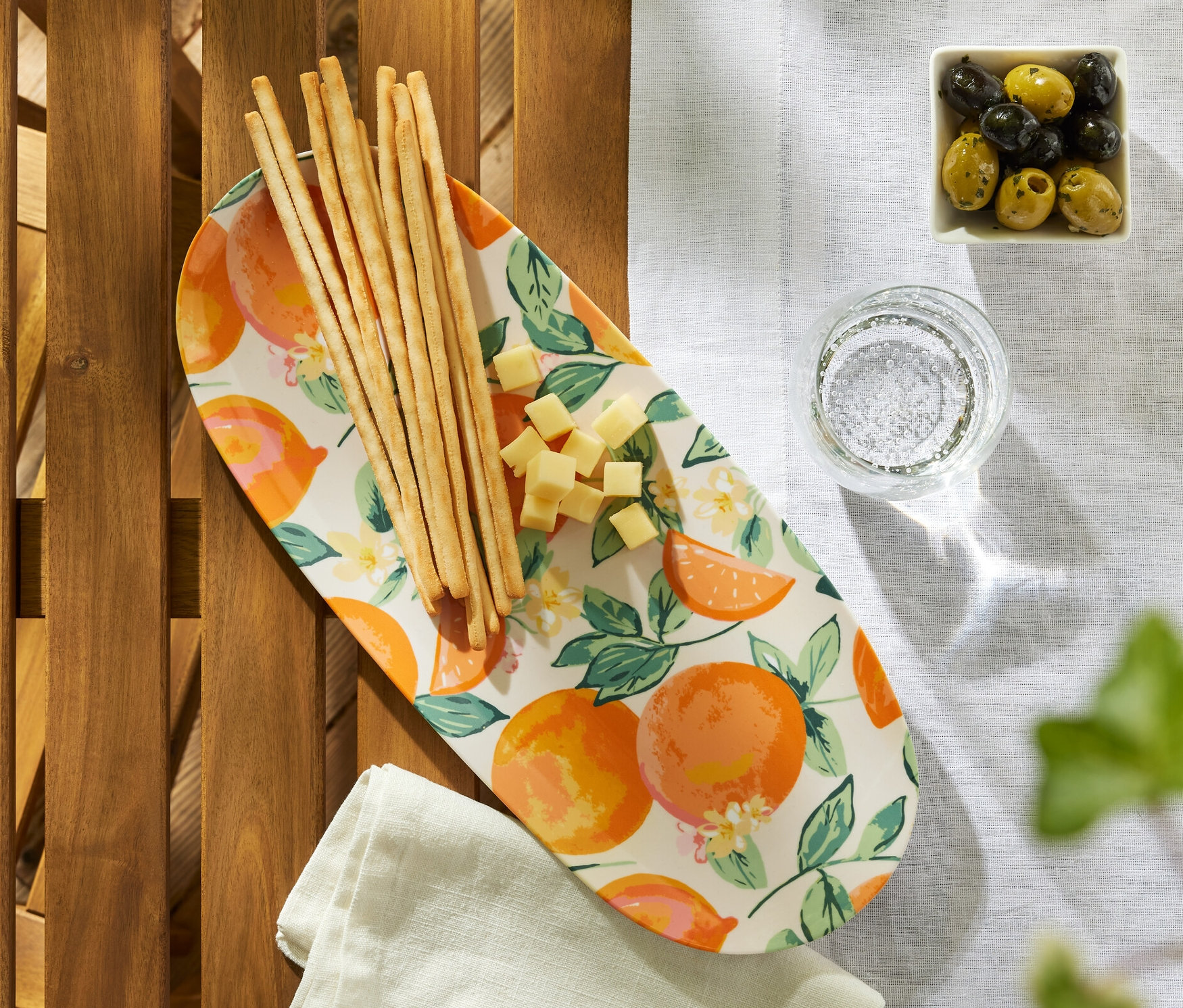 Egy fa asztalon egy kínálótálon grissini és sajt, egy pohár víz és egy tál olajbogyó található.