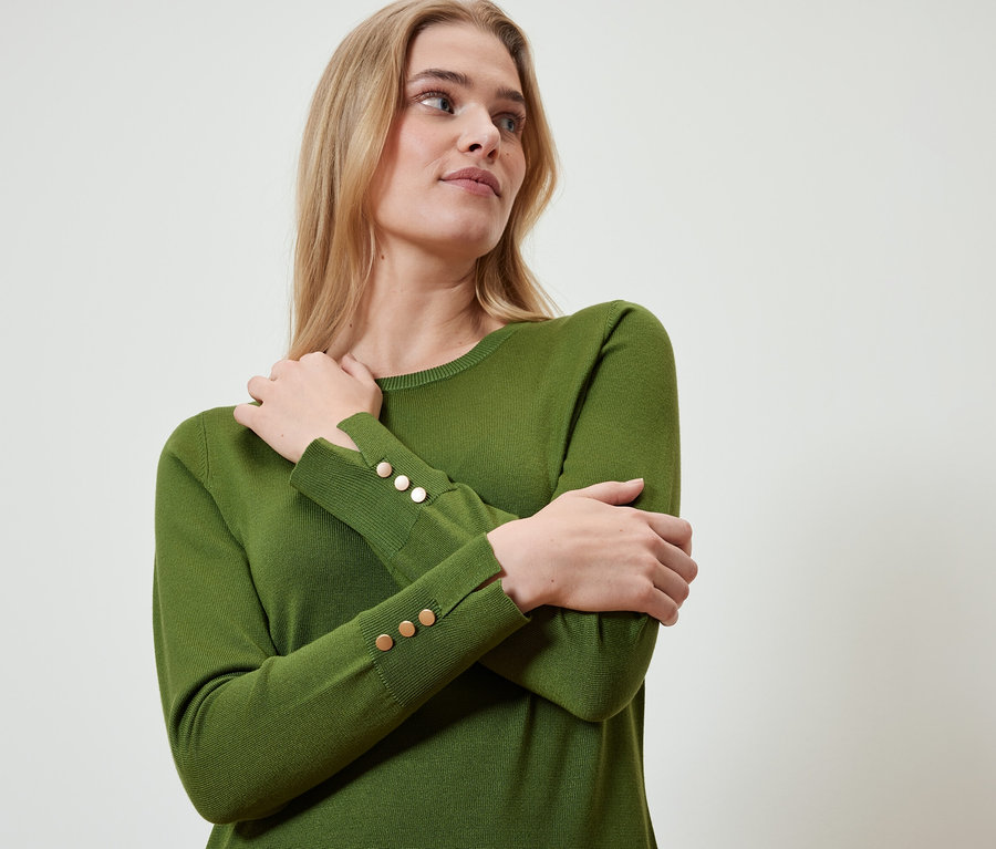 Szőke nő olívazöld pulóvert visel, arany gombokkal az ujján, karjai keresztbe vannak téve.