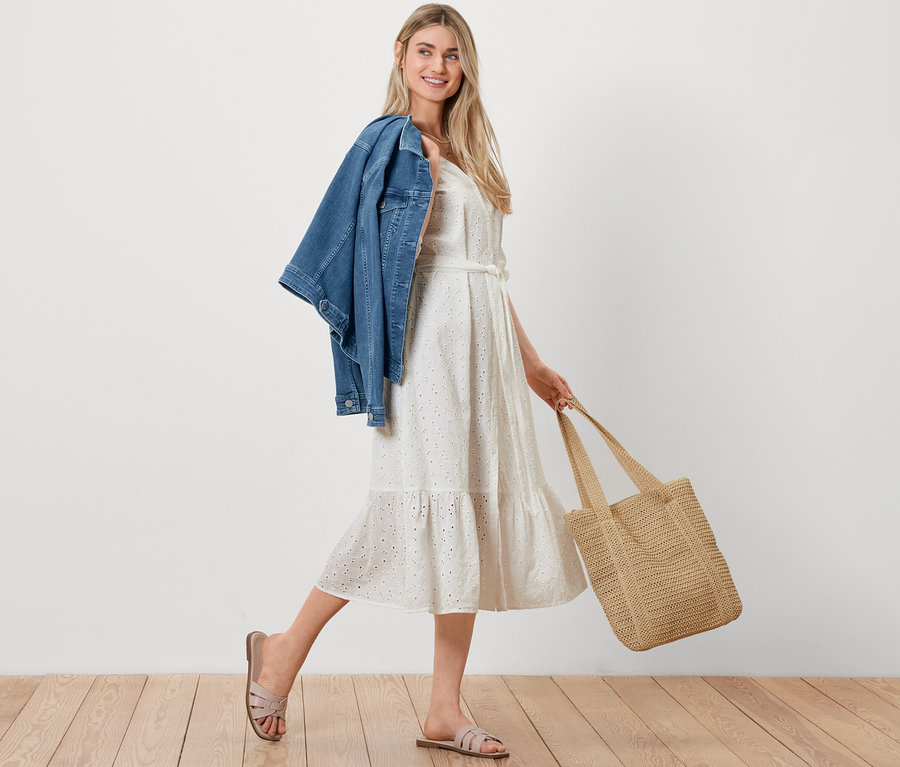 Egy nő sétál, vállán egy kék farmerdzsekivel, kezében egy háncs hatású shopper táskával és lyukhímzéses blúzruhát visel.