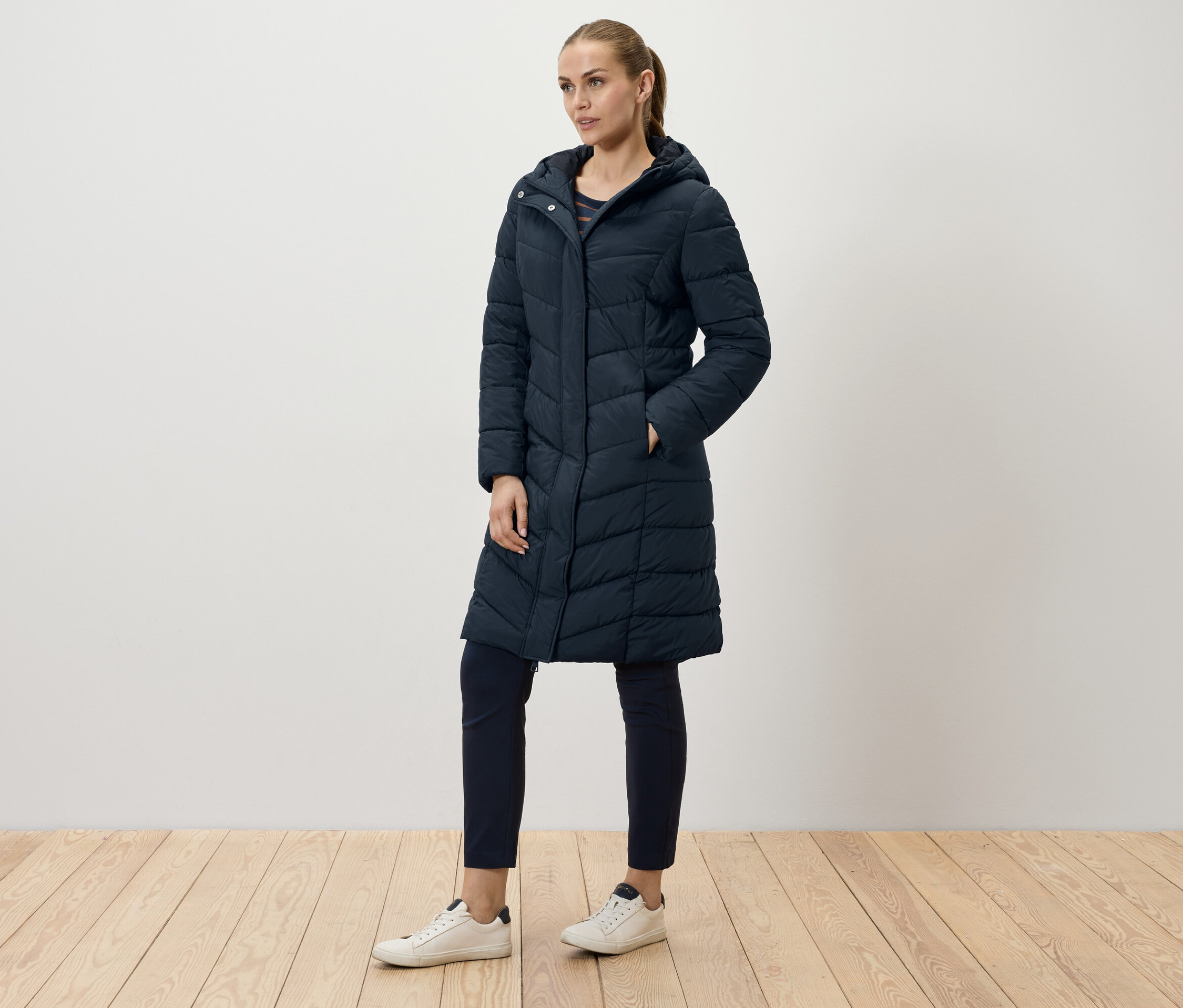 Egy nő pózol egy hosszú, sötétkék steppelt kabátban, sötétkék nadrágban és fehér tornacipőben.