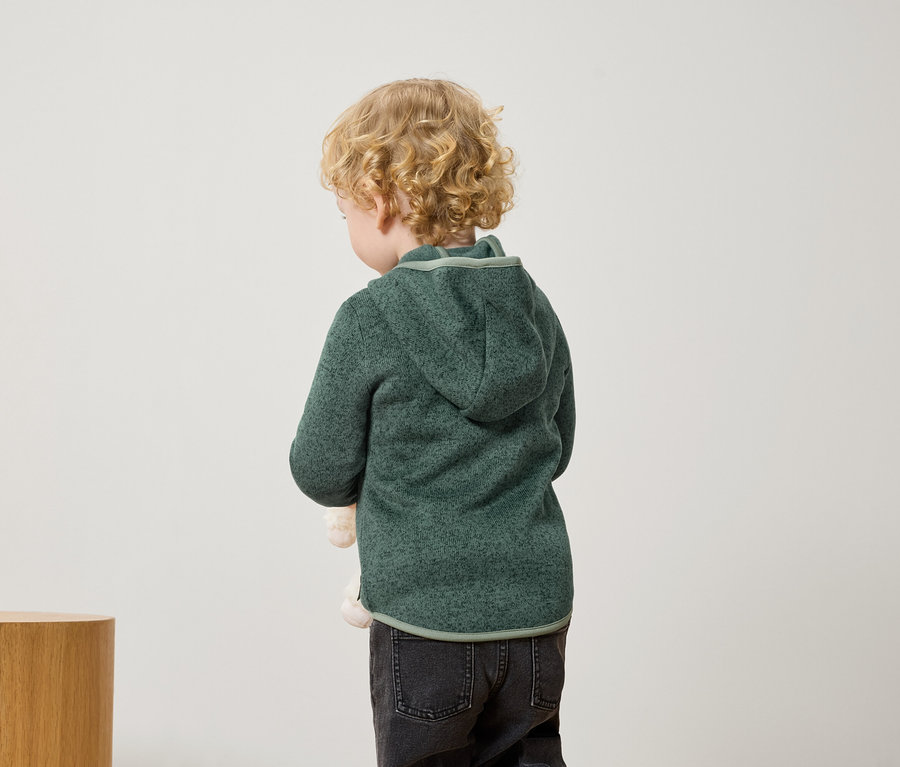 Egy szőke, göndör hajú kisfiú háttal áll, zöld kapucnis pulóvert és fekete farmert visel.
