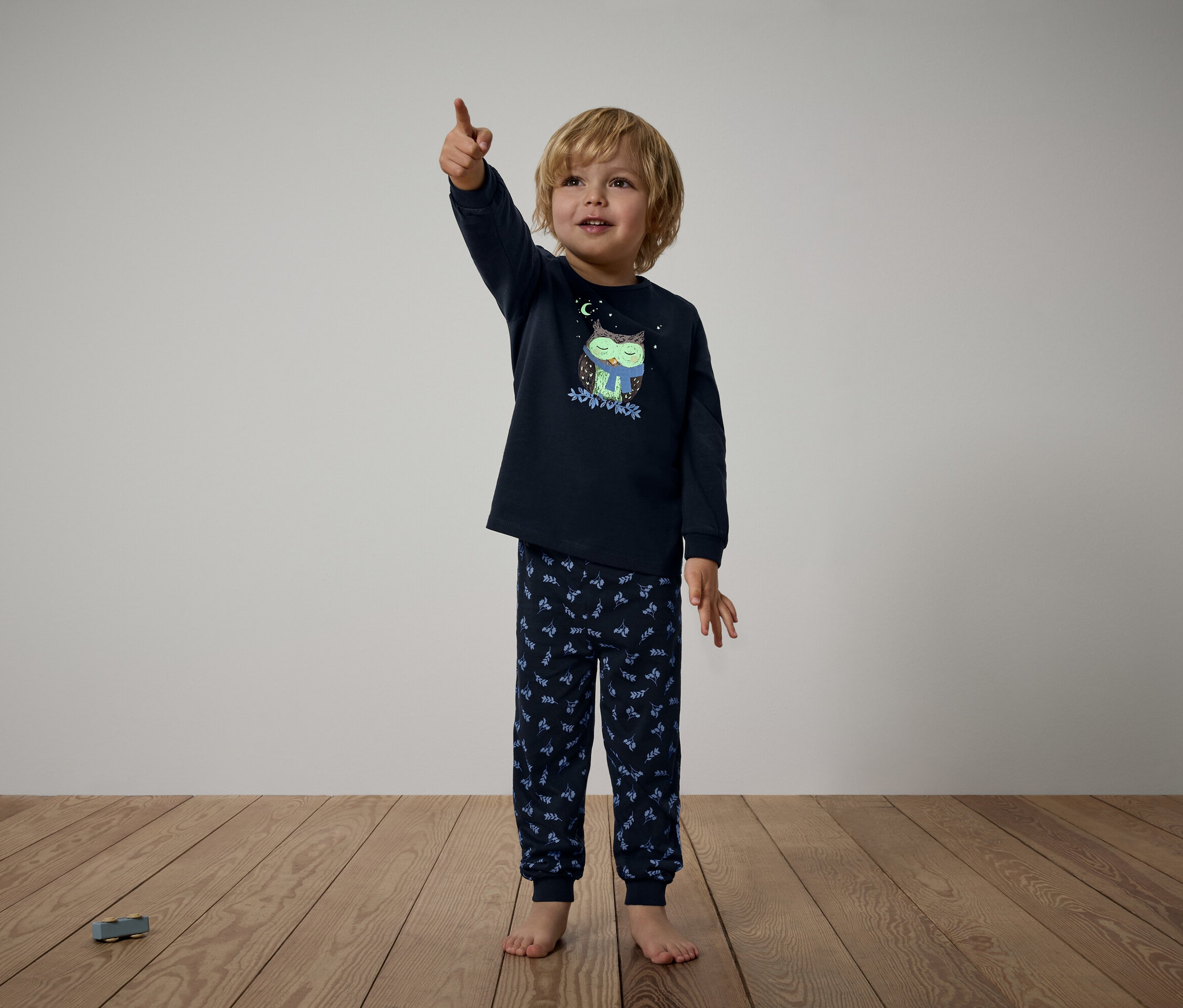 Kisfiú sötétkék, baglyos pizsamában felfelé mutat az ujjával.