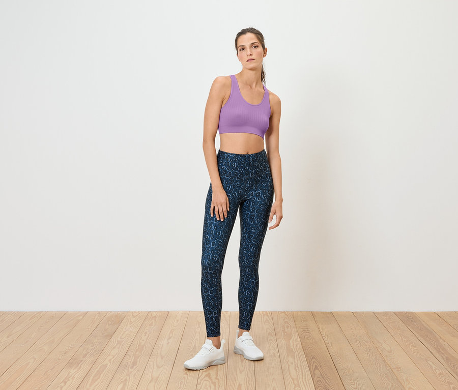 Egy nő áll a fapadlón, lila sportmelltartót és kék leggingset visel.