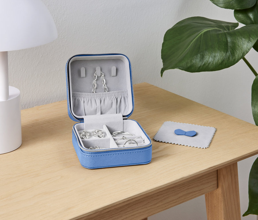 Egy nyitott, kék ékszerdoboz ezüst ékszerekkel egy fa asztalon áll egy fehér lámpa és egy zöld növény mellett.