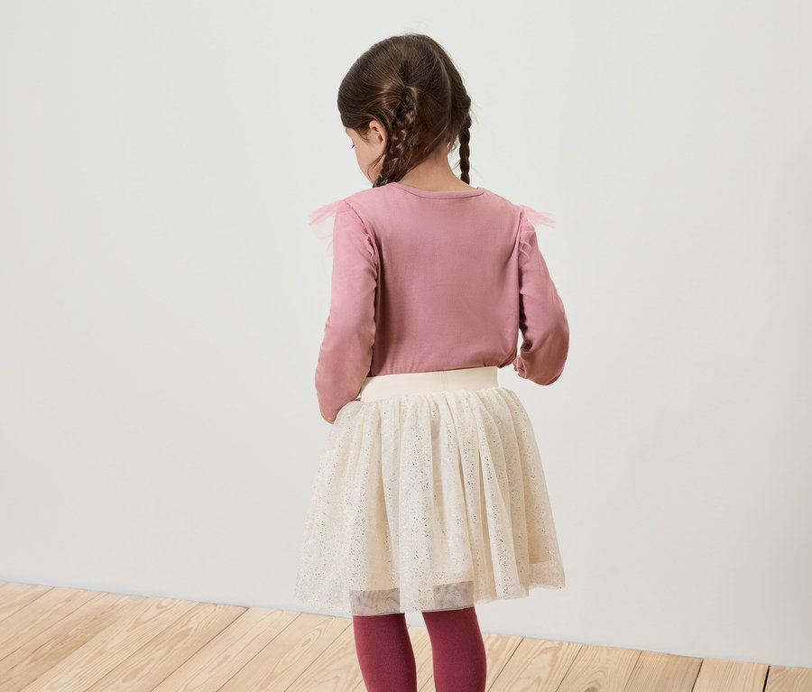 Egy hátulról látható lány rózsaszín, hosszú ujjú inget és fehér szoknyát visel.