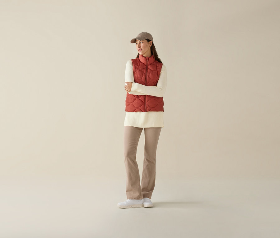 A nő keresztbe tett karokkal steppelt mellényt, krémszínű felsőt, bézs nadrágot és baseball sapkát visel.