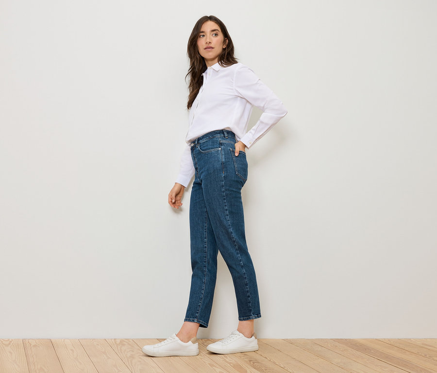 Nő pózol kék Mom Jeans – Fit »Nyla« farmerben és fehér ingben.