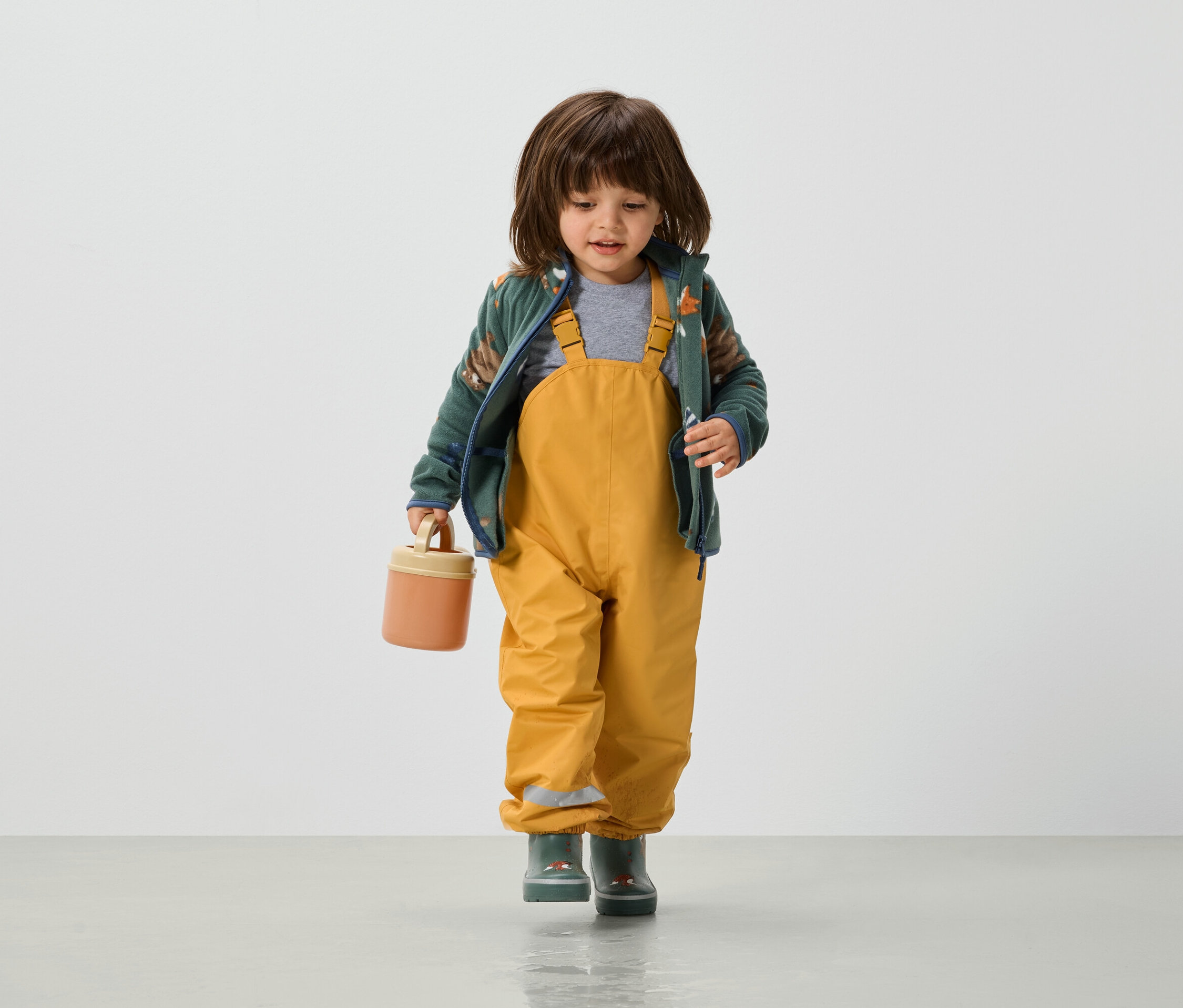 Egy kisgyerek sárga kantáros nadrágban, zöld kabátban és gumicsizmában narancssárga uzsonnásdobozt tart a kezében.