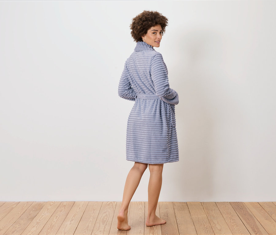 Egy nő visszanéz a válla fölött, és egy kék köntösben pózol a fapadlón.