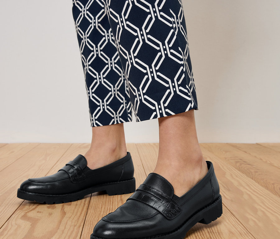 Egy személy lábai fekete mokaszinban és kék mintás nadrágban, fa padlón állva.