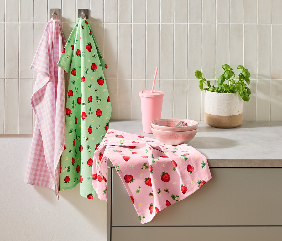 Három konyharuha, egy rózsaszín pohár szívószállal, két rózsaszín tál és egy cserép bazsalikom áll a konyhapulton.