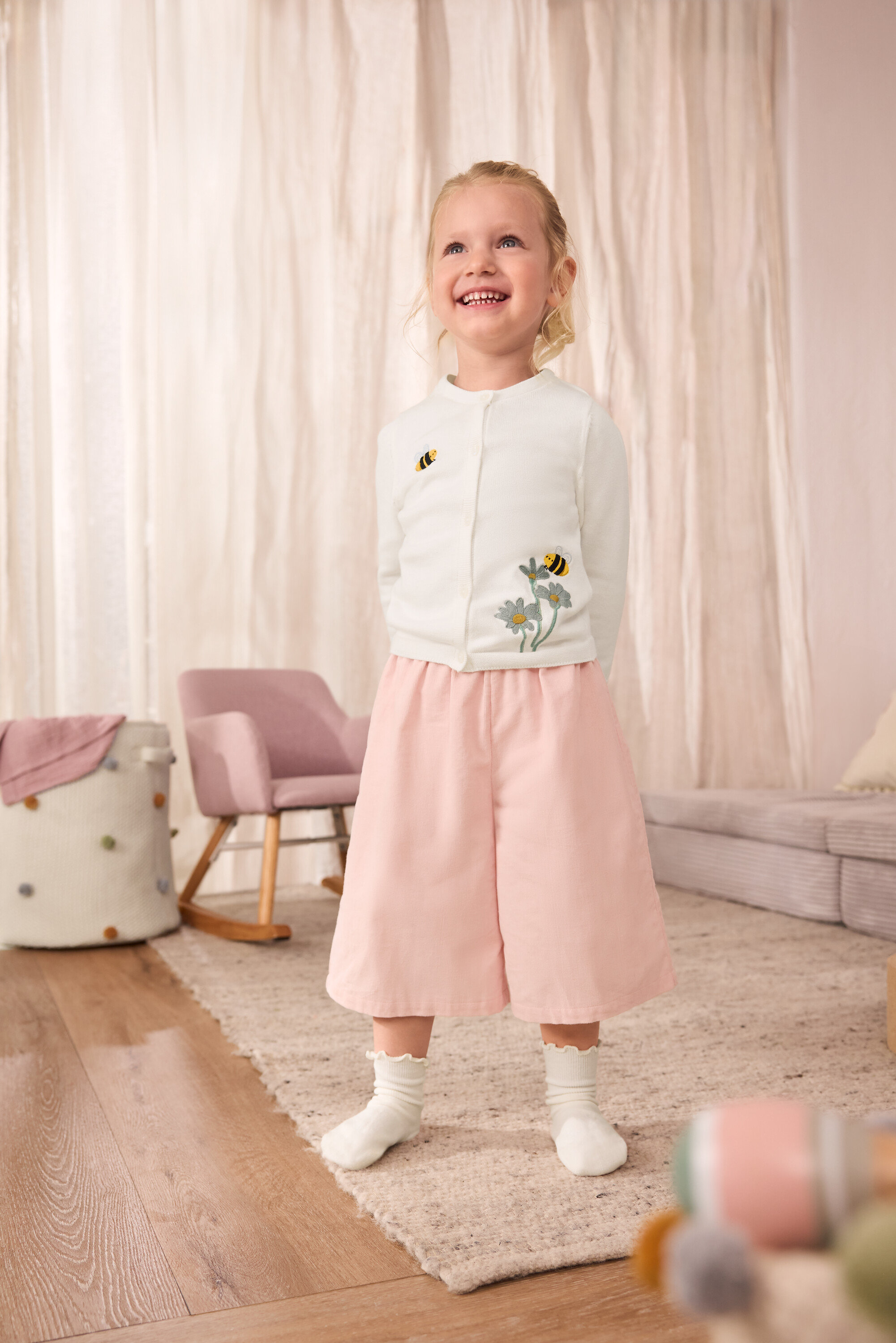 Egy mosolygó kislány hímzett méhecskés kötött kardigánt, kordbársony culotte nadrágot és 5 pár fodros zoknit visel.