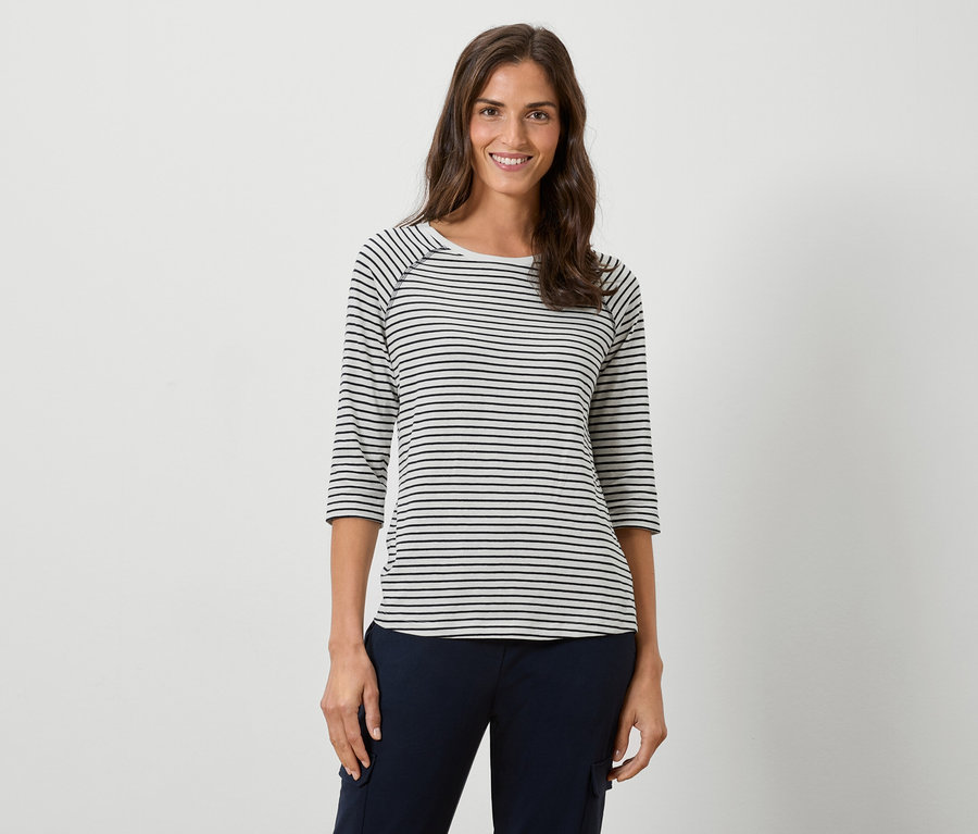 Nő visel egy csíkos, hosszú ujjú inget és egy sötétkék nadrágot fehér háttér előtt.