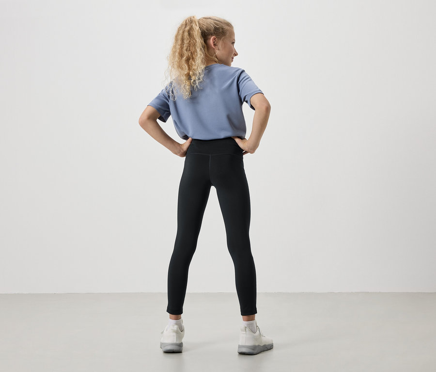 Hátulnézet egy fiatal lányról szőke, göndör hajjal, kék pólóban, fekete leggingsben és fehér tornacipőben, kezét csípőre téve.