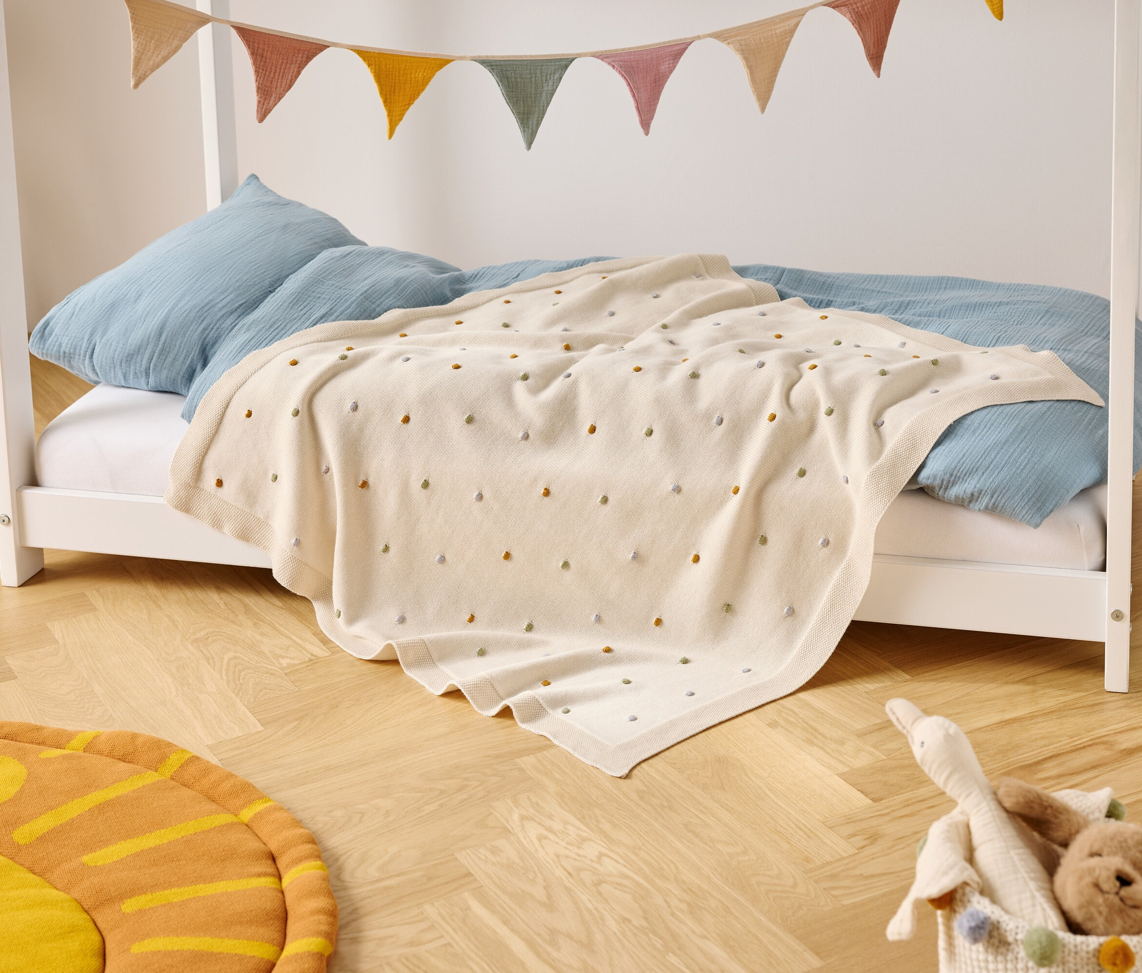 Gyerekágy kék párnákkal és színes pöttyös krémszínű takaróval áll egy fapadlón. Az ágy fölött színes zászlós szalag lóg.