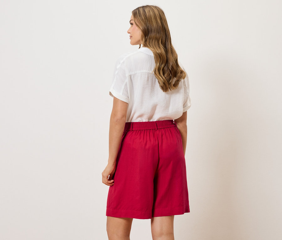 Hátulnézet egy hullámos hajú nőről, aki fehér blúzt és piros rövidnadrágot visel.