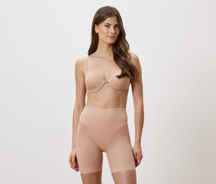 Nő visel egy testszínű melltartót és alakformáló rövidnadrágot fehér háttér előtt.
