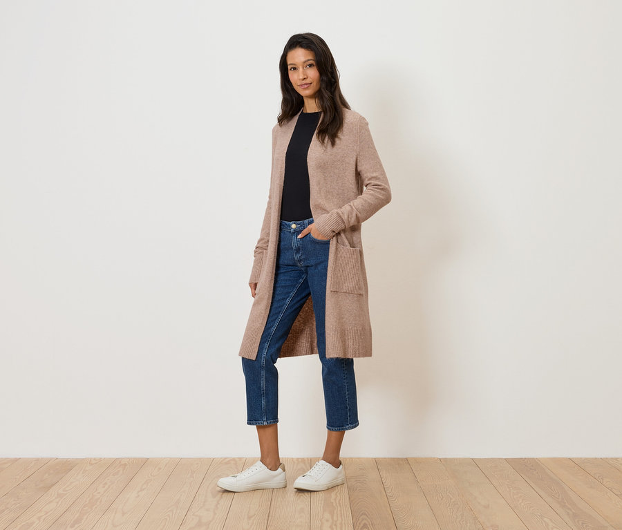 Egy nő áll egy fehér fal előtt, hosszú, bézs színű kardigánban, farmerben és fehér tornacipőben.