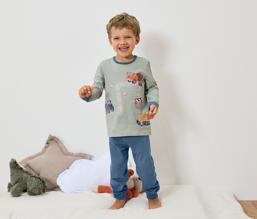 Egy mosolygó fiú mezítláb áll egy fehér felületen, szürke pizsamát visel kék mandzsettákkal, és egy kotrógép és betonkeverő motívummal.