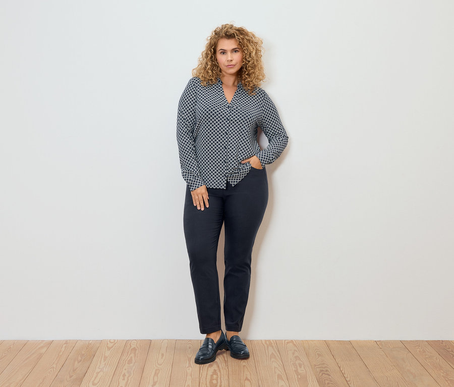 Nő mintás blúzban és bengalin nadrágban pózol egy fal előtt.