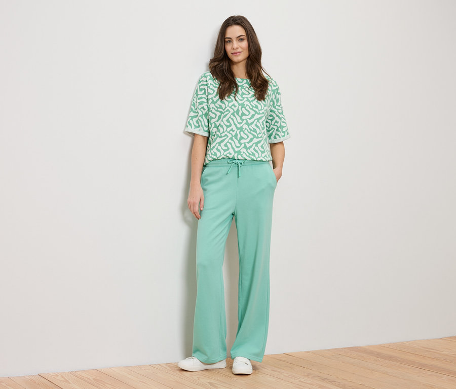 Egy nő a falnak támaszkodik, zöld mintás felsőt és zöld nadrágot visel.