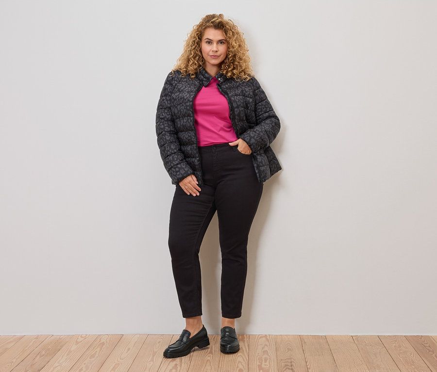 Göndör hajú nő szürke-fekete, steppelt dzsekit visel egy rózsaszín felső felett, fekete farmerrel és loaferekkel kombinálva.