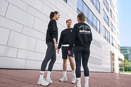 Három ember, két nő és egy férfi, állva mosolyogva beszélget. Mindannyian fekete szabadidőfelsőt és fehér Uniszex Coffe Crew teniszzoknit viselnek. A férfi fekete sport rövidnadrágot visel. Az egyik nő antracit színű, leopárdmintás női sportnadrágot, a másik fekete női sportnadrágot visel.
