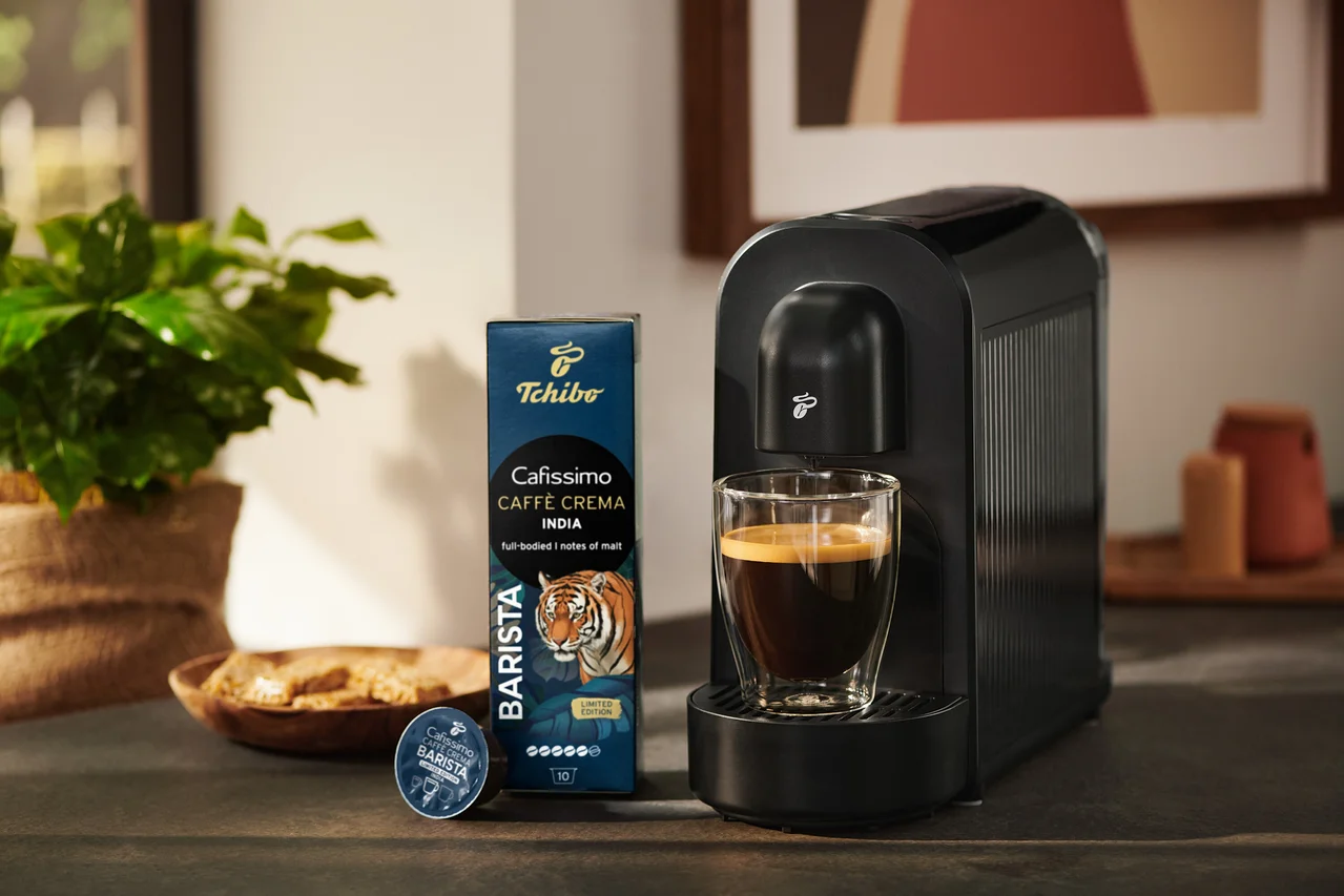 Fekete Tchibo Cafissimo kávéfőző egy pohár kávéval, mellette egy csomag Tchibo Cafissimo Caffè Crema India kávékapszula és egyetlen kapszula.