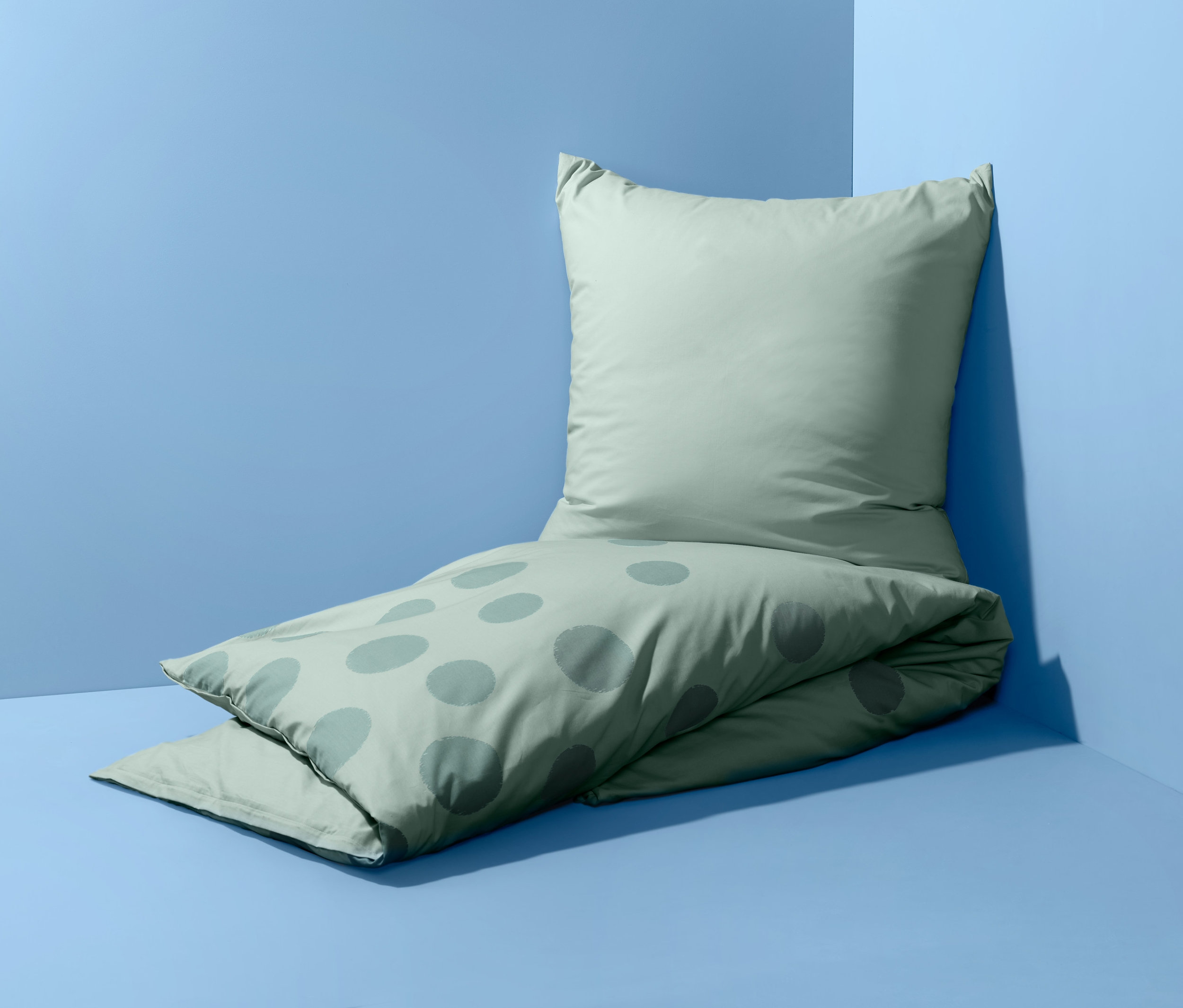 Világoszöld, pöttyös, kétoldalas perkál ágynemű, egyszemélyes méretben a fal mellett a földön.