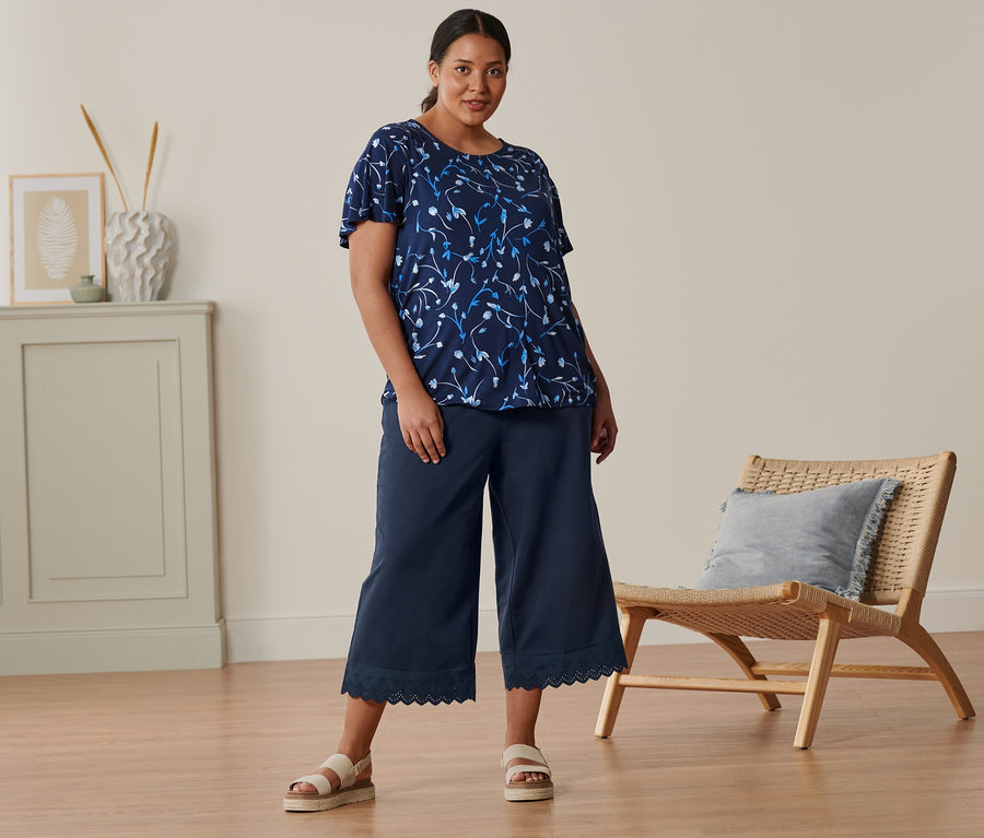 Egy nő áll, sötétkék virágos pólóblúzt, 3/4-es bőszárú nadrágot és bézs bőr szandált visel.