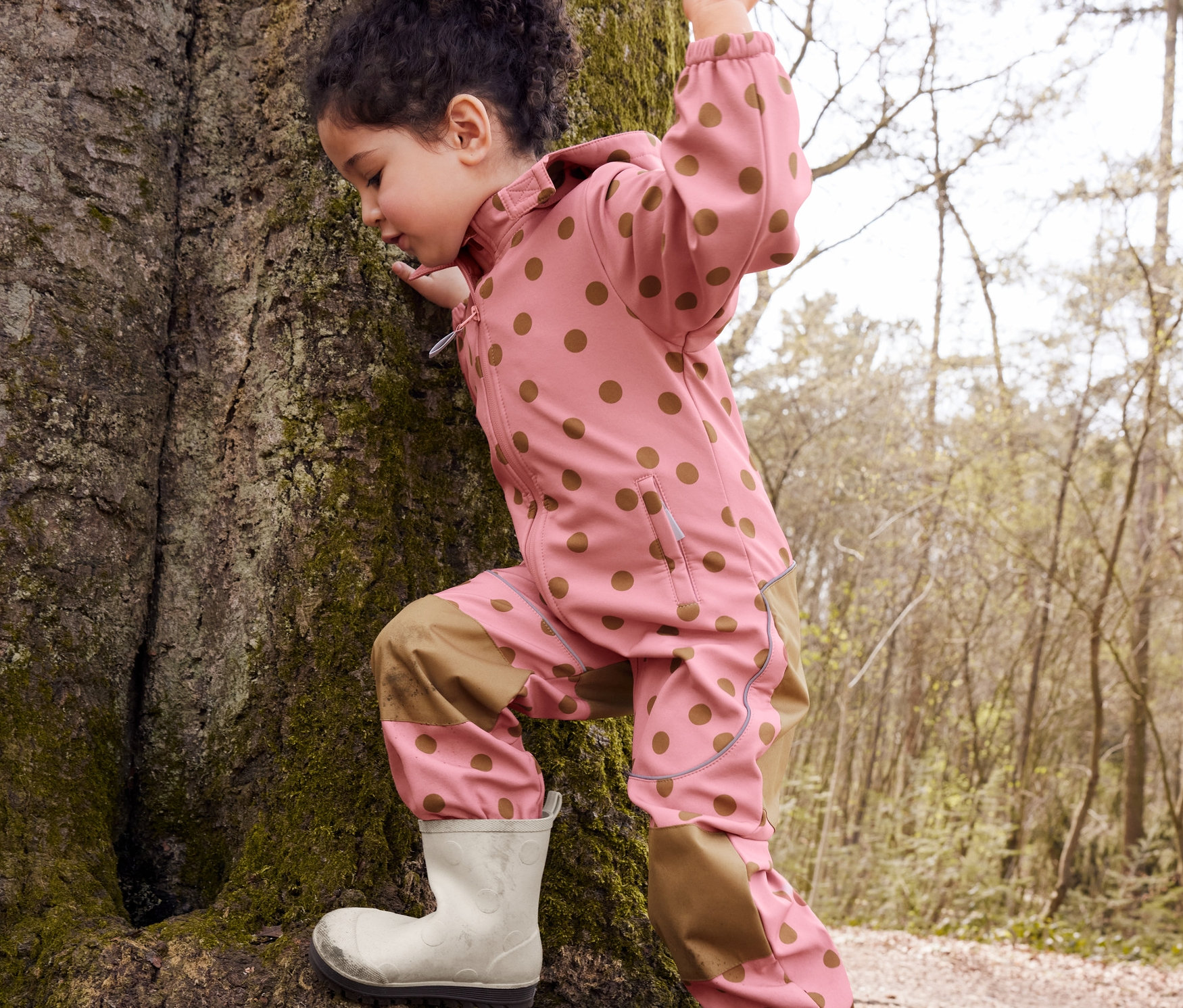 Egy kislány pöttyös softshell kezeslábasban és gumicsizmában fára mászik.
