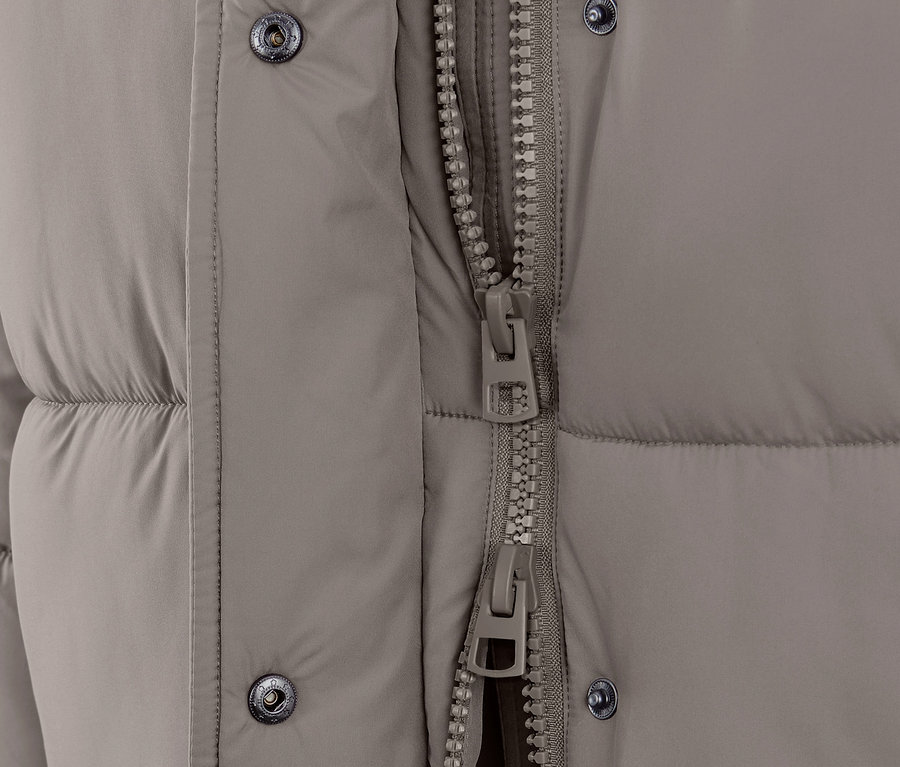 Egy taupe színű, steppelt kabát részlete kapucnival és cipzárral.