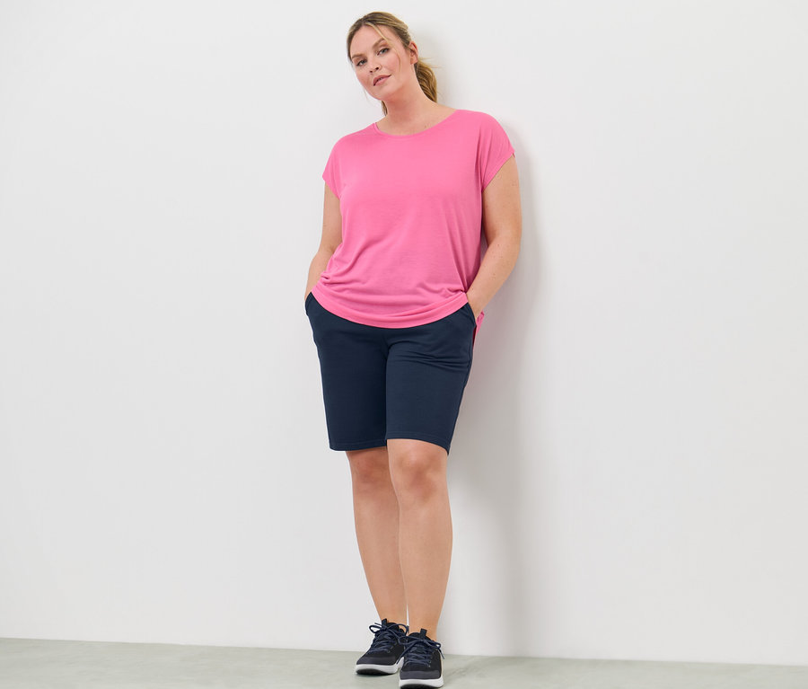 Nő áll, kezével a rövidnadrágja zsebében. Egy rózsaszín sportfelsőt visel.