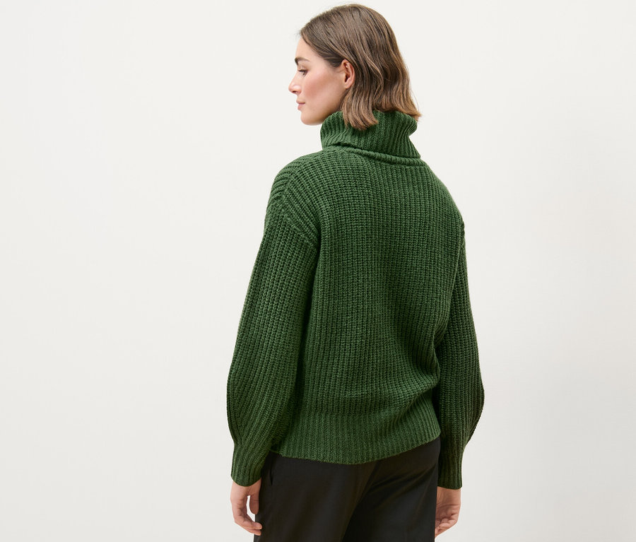 Nő visel sötétzöld, gyapjúval készült garbónyakú pulóvert és fekete nadrágot.