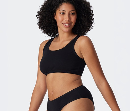 Mosolygó, sötét, göndör hajú nő fekete SCHIESSER bustier melltartót és fekete alsóneműt visel.