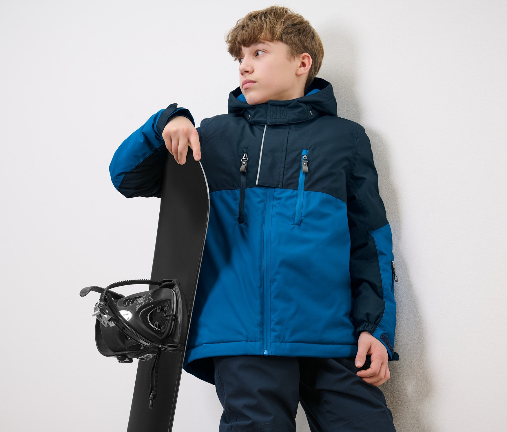 Egy fiú fiú síkabátban áll egy snowboard mellett.