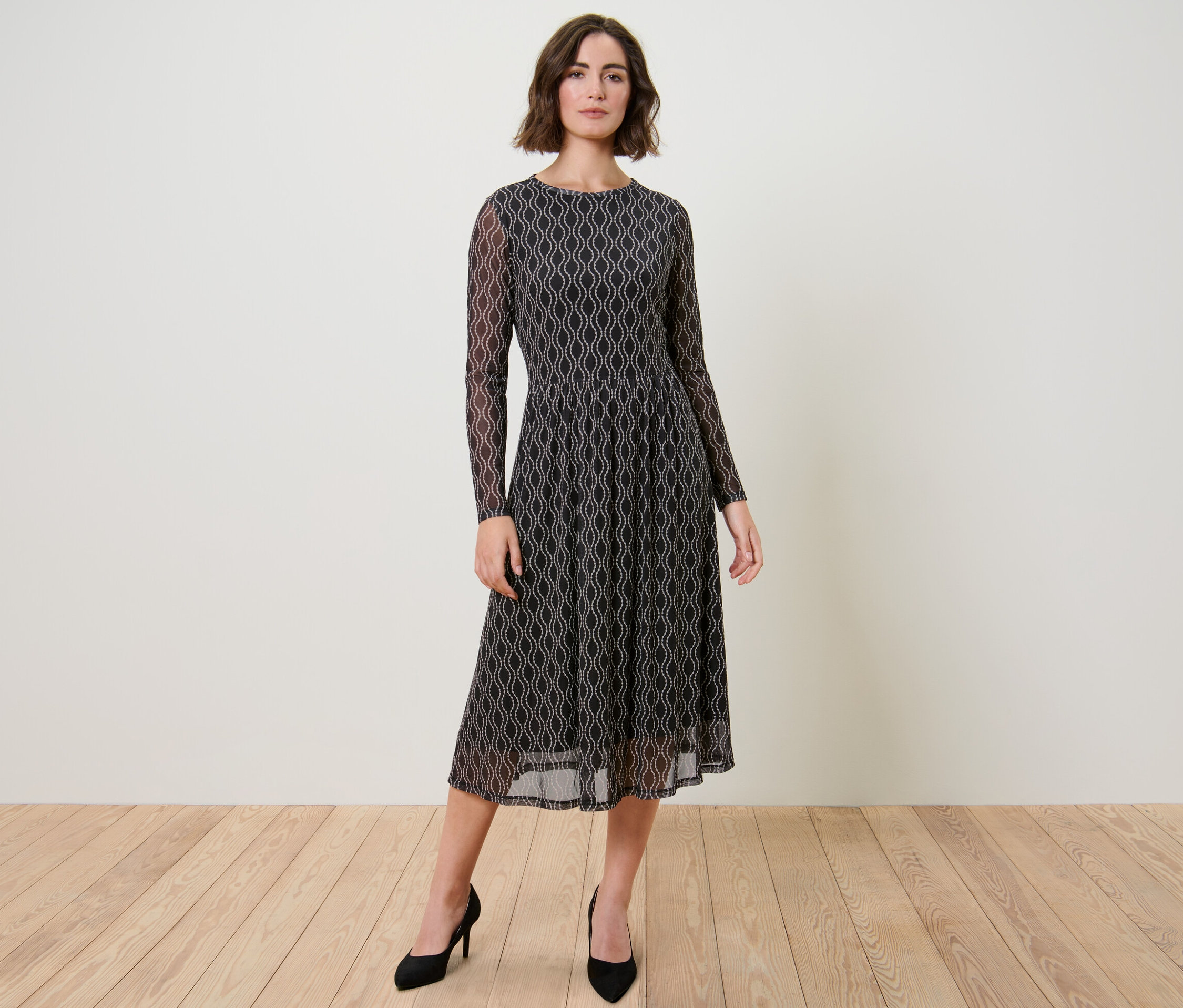 Nő pózol mintás, fekete mesh midiruhában és fekete magassarkúban.
