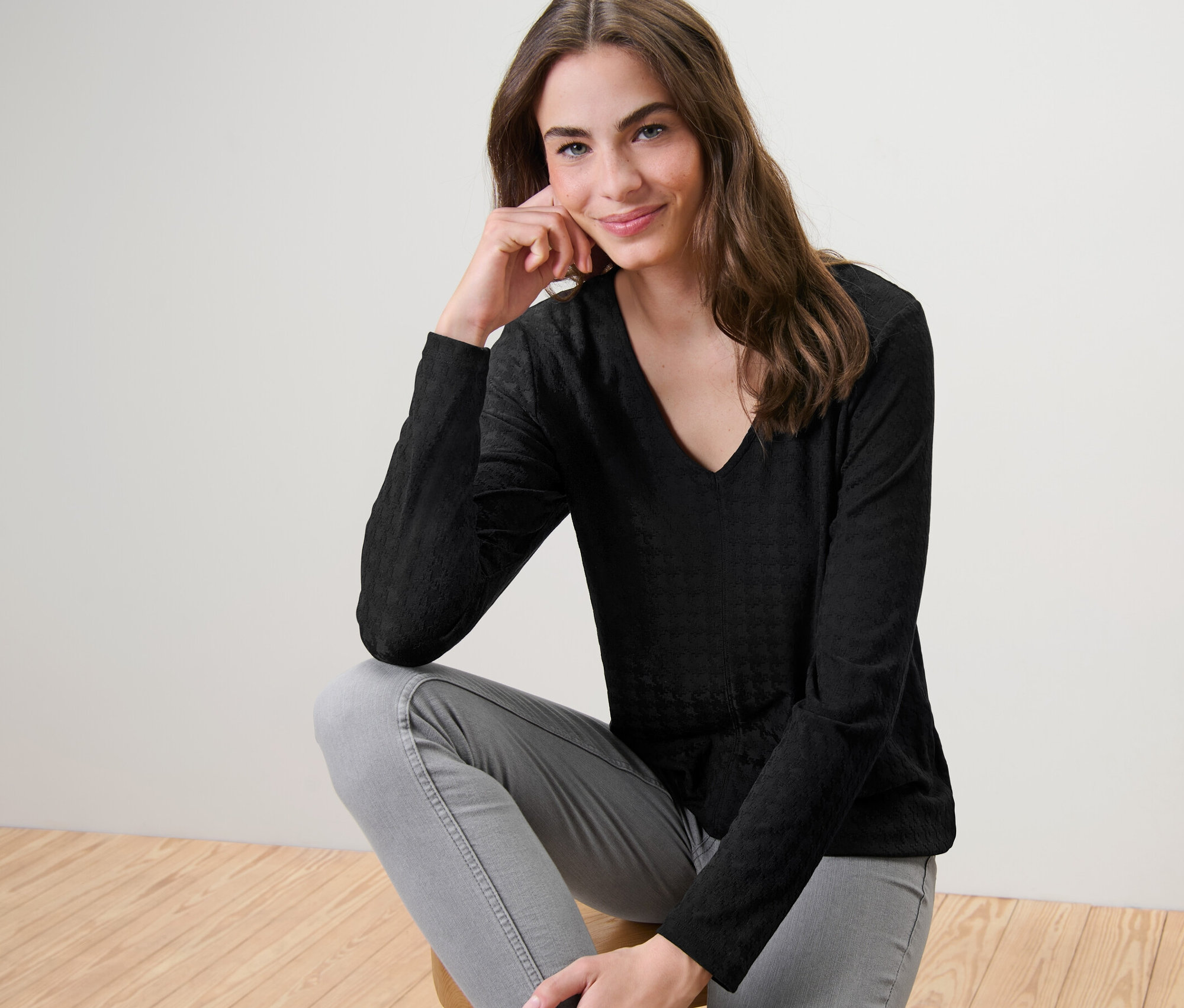 Egy nő ül egy széken, és fekete, strukturált, hosszú ujjú pulóvert visel.