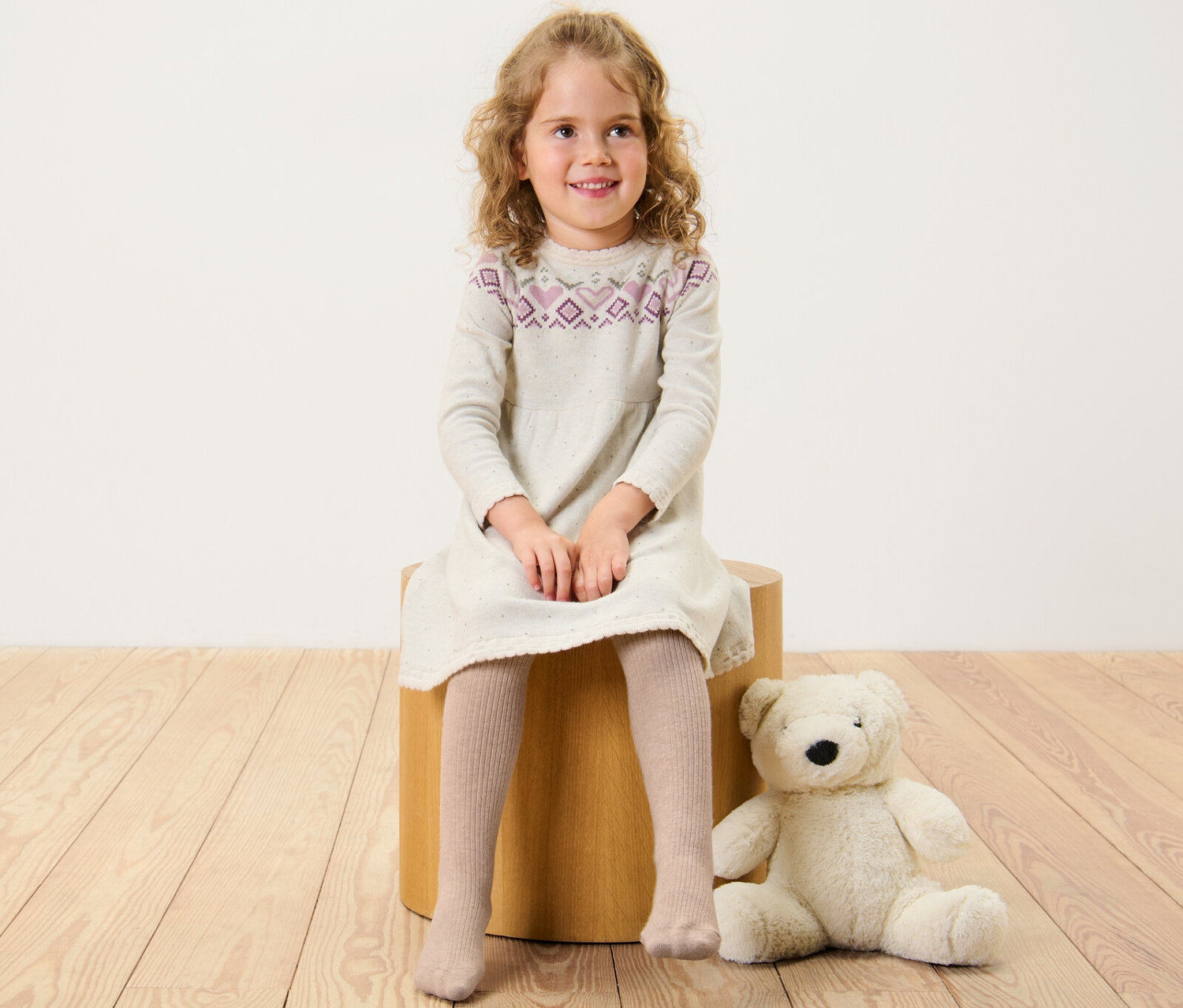 Egy kislány ül egy fa ülőkén, ruhát és Kisgyerek harisnyát visel gyapjúval, bézs.