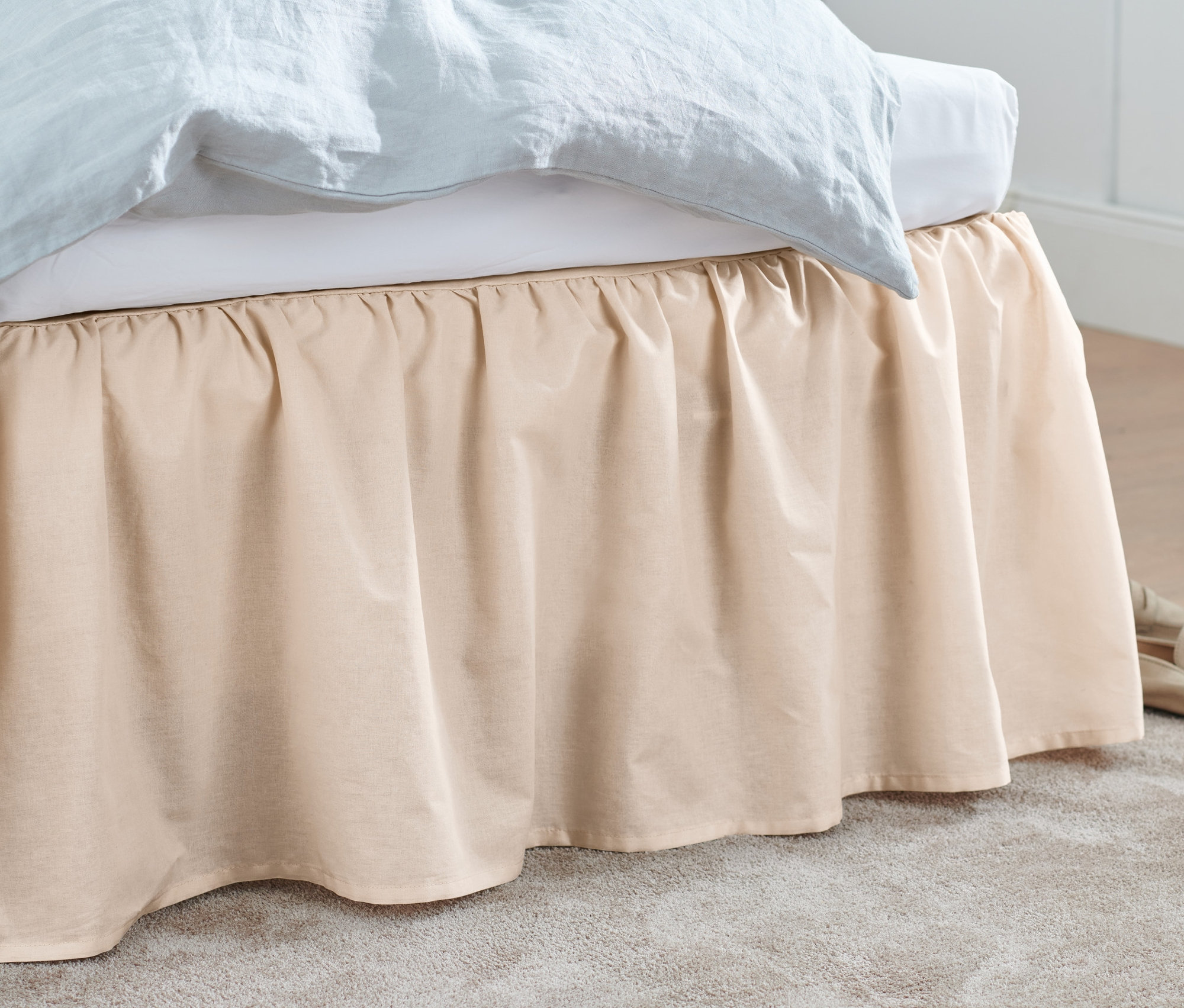 Egy bézs ágytakaró takar egy ágyat. Bézs színű cipő és egy szürke szőnyeg van a padlón.