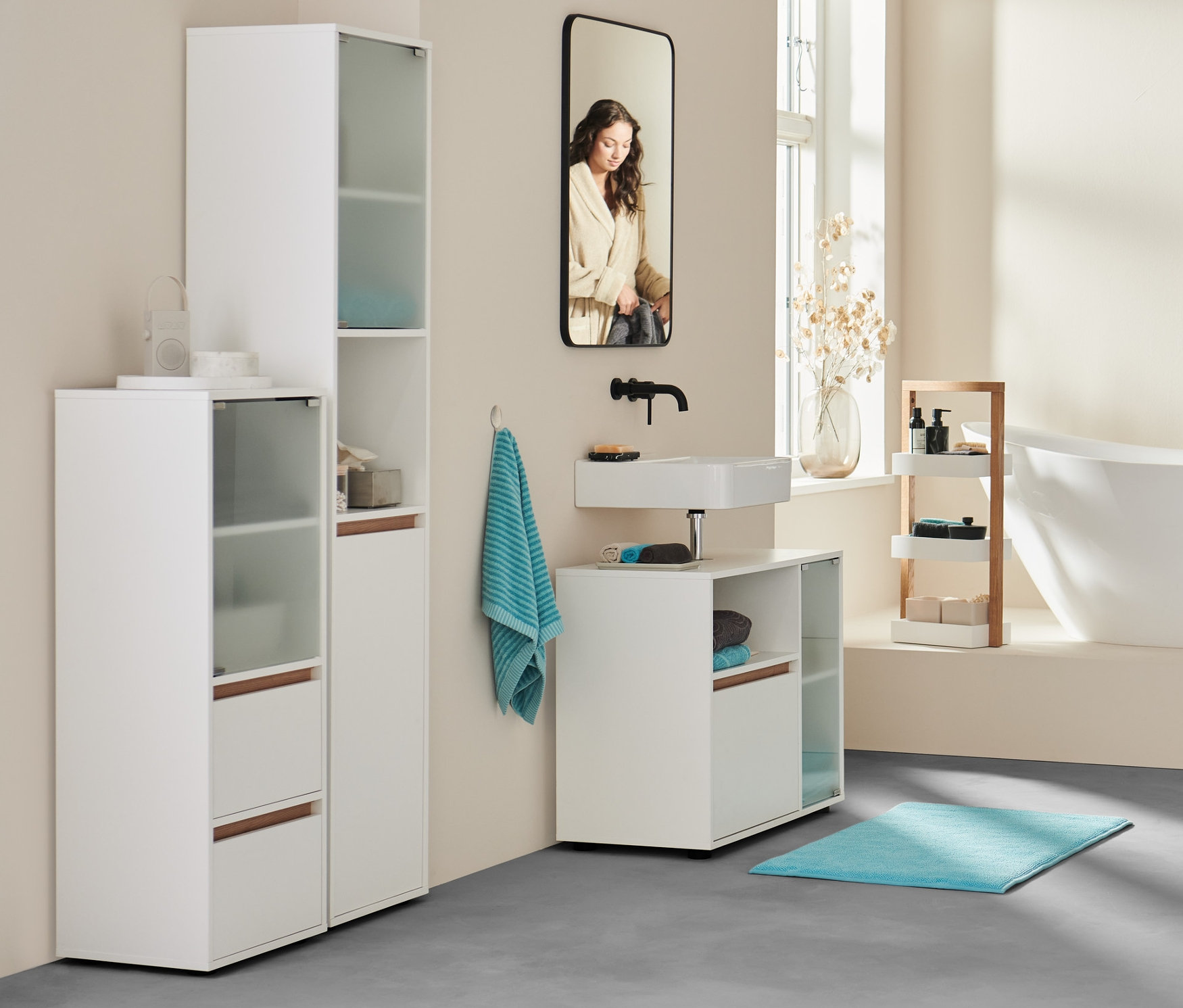 A fürdőszobában egy nő áll fürdőköpenyben a fürdőkád mellett. Előtérben fürdőszobai szekrény, extranagy antracit fürdőlepedő és 2 vízkék törölköző.