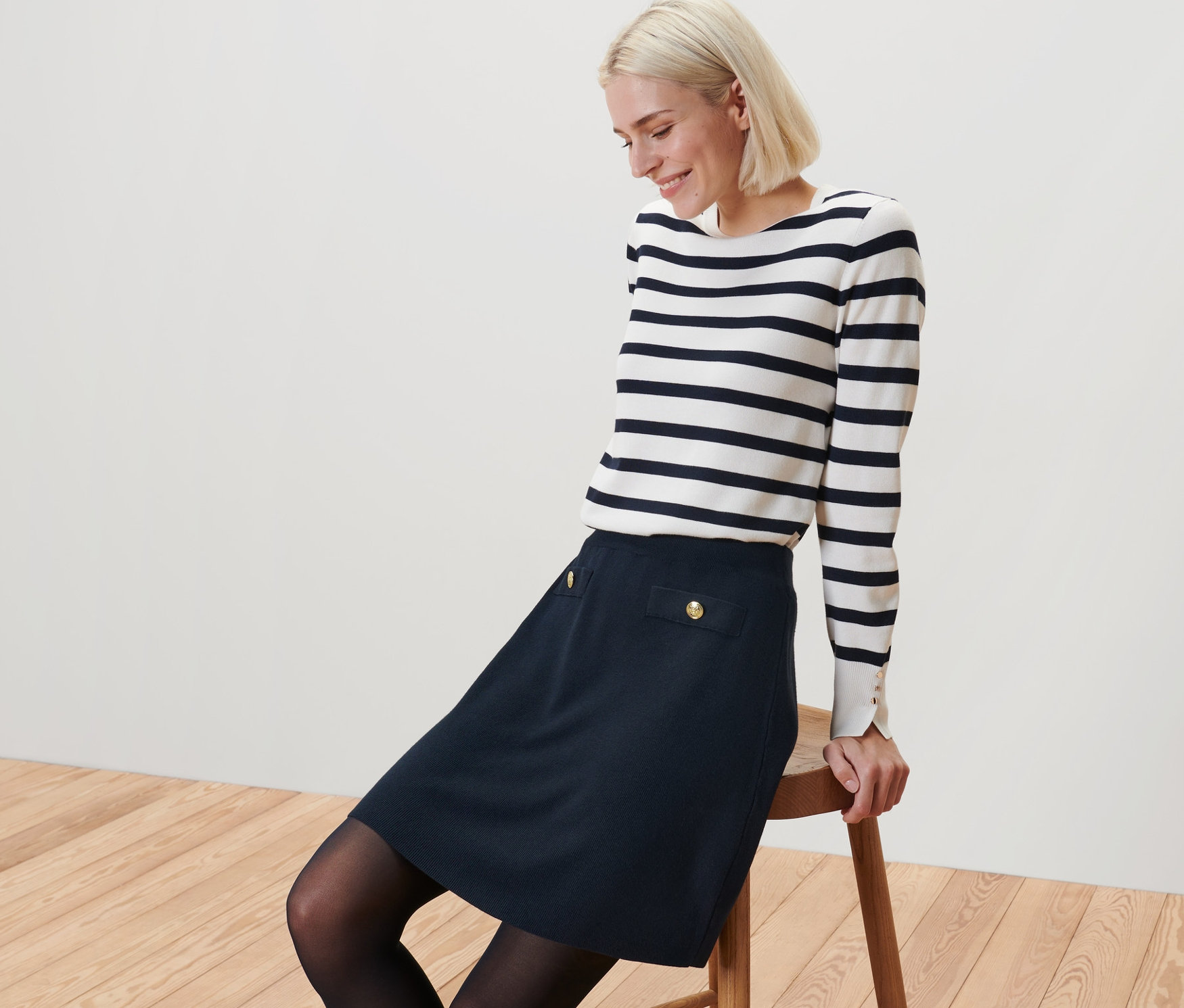 Szőke nő ül egy széken, díszgombos kötött szoknyát és csíkos felsőt visel.