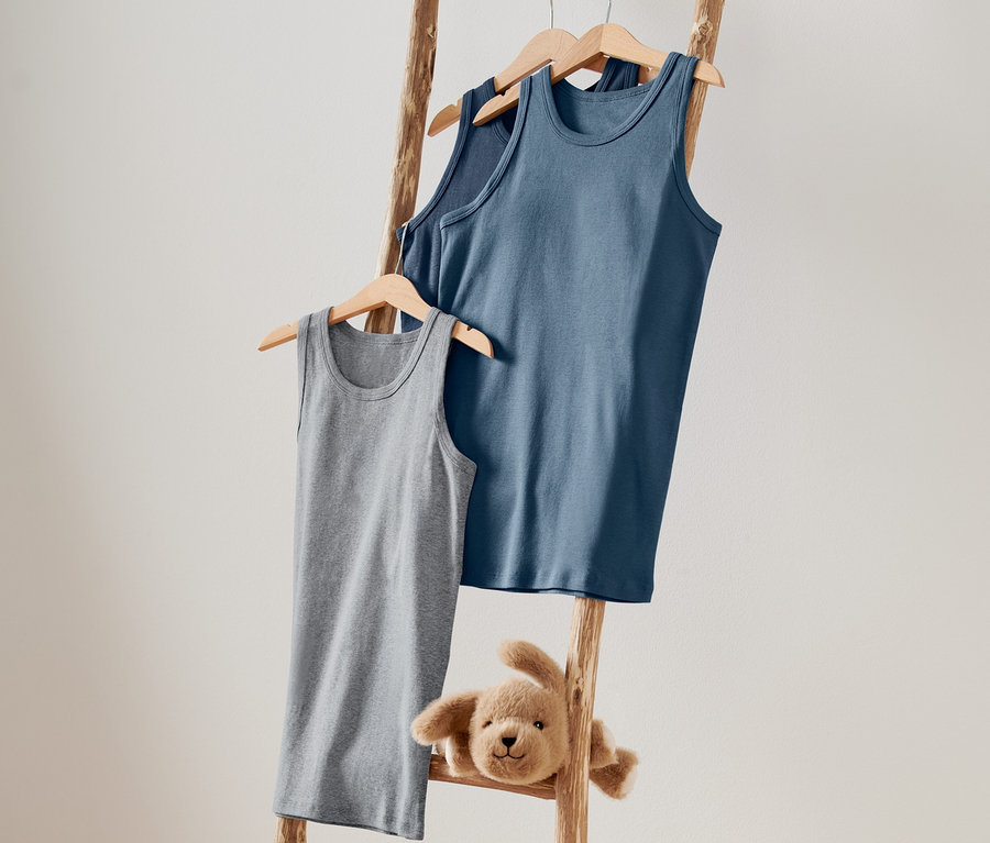 Egy falnak támasztott fa létrán szürke és kék fiú trikók lógnak vállfán, valamint egy plüss kutyus.