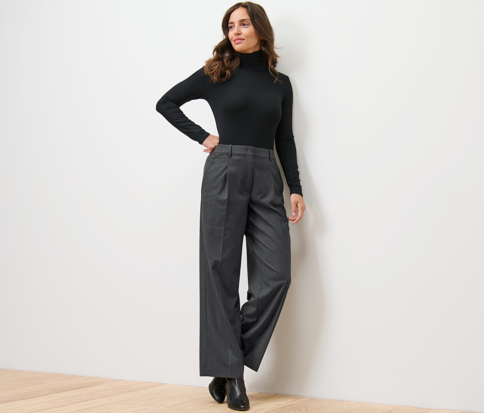 Nő pózol fekete garbóban és szürke női szövött nadrágban a falnak támaszkodva.