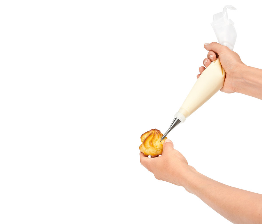 Egy kéz egy habzsák segítségével krémmel tölt meg egy péksüteményt.