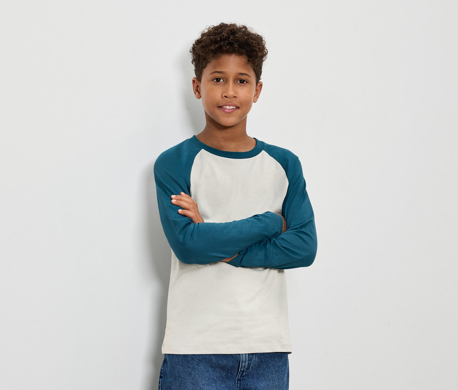 Barna, göndör hajú fiú áll keresztbe tett karokkal, kékeszöld hosszú ujjú inget és farmert visel.